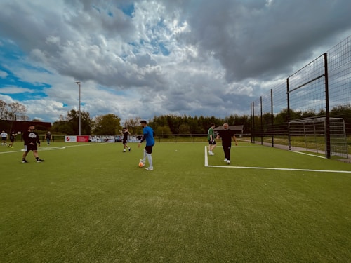Several people are playing football on an outdoor artificial turf field surrounded by a wire fence. The sky above is overcast with patches of blue visible through the clouds. There are advertising boards along the side of the field and trees and shrubbery in the background.