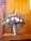 A floral arrangement consisting of white and purple flowers, including daisies and carnations, is elegantly displayed in an ornate silver vase. The bouquet is set against a backdrop of warm, wooden paneling which enhances the vibrancy of the flowers. Green foliage adds fullness to the arrangement.