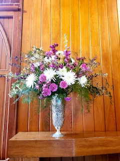 A floral arrangement consisting of white and purple flowers, including daisies and carnations, is elegantly displayed in an ornate silver vase. The bouquet is set against a backdrop of warm, wooden paneling which enhances the vibrancy of the flowers. Green foliage adds fullness to the arrangement.