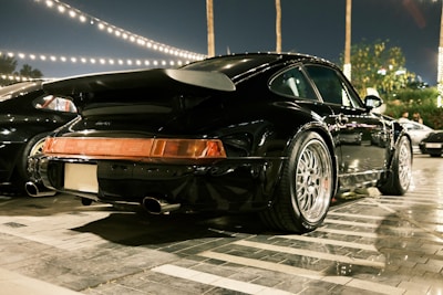 A sleek black sports car is parked under string lights, reflecting the ambient glow on its polished surface. The vehicle's aggressive rear wing and distinctive wheel design hint at its performance capabilities. Nearby, other cars are similarly parked, suggesting a gathering of high-performance vehicles.