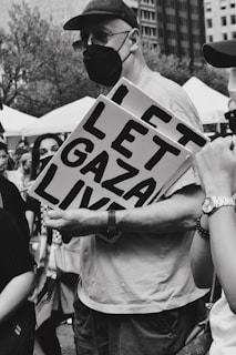 A black and white photograph of a protest scene with a man holding a sign that reads 'LET GAZA LIVE'. The man is wearing a face mask and a cap. People are gathered around, and the background shows trees and buildings.