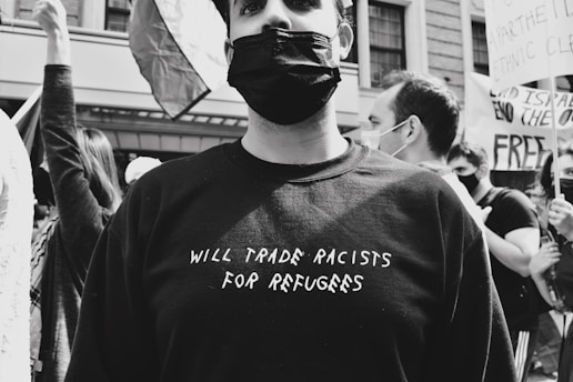 A group of people are participating in a protest or demonstration. The individual in the foreground is wearing a black mask and a black shirt with the phrase 'WILL TRADE RACISTS FOR REFUGEES' written on it. Other people in the background are holding signs and one of them is raising their arm. The mood is serious, and the setting appears to be urban, indicated by the building in the background.