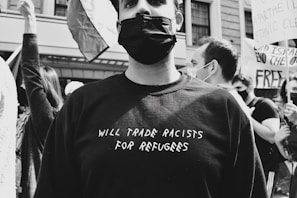 A group of people are participating in a protest or demonstration. The individual in the foreground is wearing a black mask and a black shirt with the phrase 'WILL TRADE RACISTS FOR REFUGEES' written on it. Other people in the background are holding signs and one of them is raising their arm. The mood is serious, and the setting appears to be urban, indicated by the building in the background.