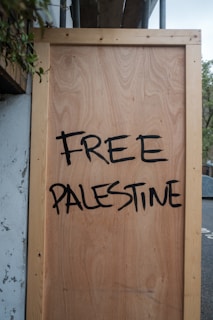 A large wooden board with the words 'Free Palestine' painted in bold, black letters. The board is propped against a wall and surrounded by some foliage in the upper left corner.
