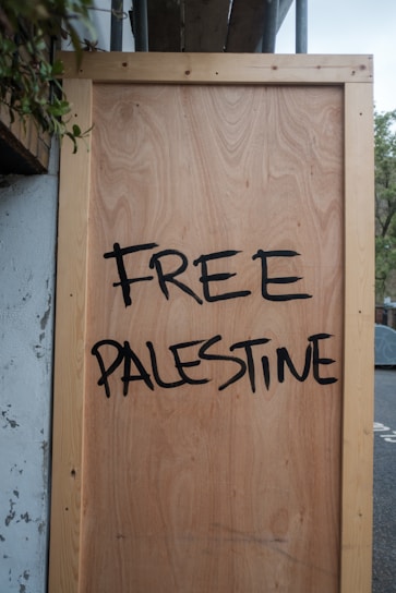 A large wooden board with the words 'Free Palestine' painted in bold, black letters. The board is propped against a wall and surrounded by some foliage in the upper left corner.