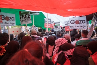 A large crowd is gathered, holding signs with messages such as 'Free Palestine' and 'End the siege.' The crowd is diverse, with people wearing a variety of clothing including headscarves and jackets. A large Palestinian flag is held overhead, creating a canopy of red, green, black, and white. The signs have text in both English and Arabic.