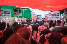 A large crowd is gathered, holding signs with messages such as 'Free Palestine' and 'End the siege.' The crowd is diverse, with people wearing a variety of clothing including headscarves and jackets. A large Palestinian flag is held overhead, creating a canopy of red, green, black, and white. The signs have text in both English and Arabic.