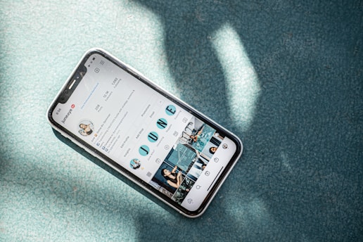 A smartphone displaying an Instagram profile screen lies on a textured teal surface with sunlight casting dappled shadows over the scene. The phone case appears clear with a slight reflection.