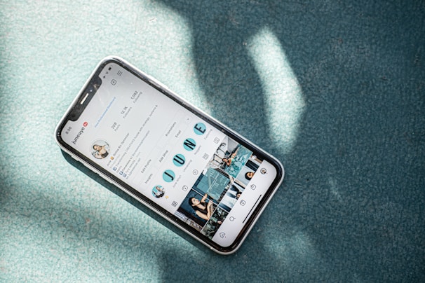 A smartphone displaying an Instagram profile screen lies on a textured teal surface with sunlight casting dappled shadows over the scene. The phone case appears clear with a slight reflection.