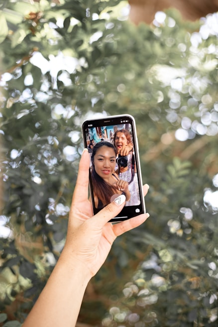 A hand with a geometric nail design holds a smartphone displaying an image of two smiling individuals, one holding a camera. The background is filled with lush green foliage and bokeh light effects, creating a vibrant and lively atmosphere.