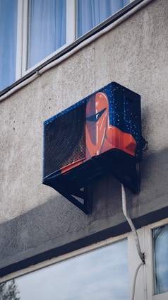 An air conditioning unit is mounted to the exterior of a building, creatively painted with artwork resembling a futuristic helmet. The building's textured concrete wall and nearby window are visible.