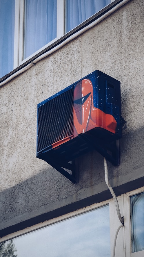 An air conditioning unit is mounted to the exterior of a building, creatively painted with artwork resembling a futuristic helmet. The building's textured concrete wall and nearby window are visible.