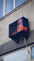 An air conditioning unit is mounted to the exterior of a building, creatively painted with artwork resembling a futuristic helmet. The building's textured concrete wall and nearby window are visible.