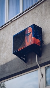 An air conditioning unit is mounted to the exterior of a building, creatively painted with artwork resembling a futuristic helmet. The building's textured concrete wall and nearby window are visible.