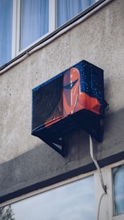 An air conditioning unit is mounted to the exterior of a building, creatively painted with artwork resembling a futuristic helmet. The building's textured concrete wall and nearby window are visible.