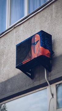 An air conditioning unit is mounted to the exterior of a building, creatively painted with artwork resembling a futuristic helmet. The building's textured concrete wall and nearby window are visible.