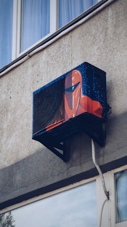 An air conditioning unit is mounted to the exterior of a building, creatively painted with artwork resembling a futuristic helmet. The building's textured concrete wall and nearby window are visible.