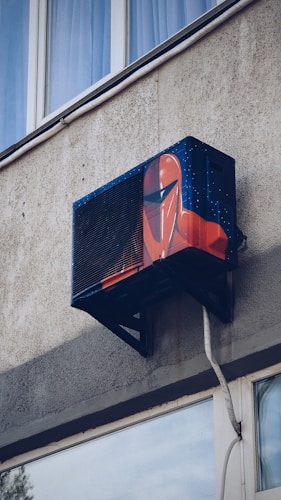 An air conditioning unit is mounted to the exterior of a building, creatively painted with artwork resembling a futuristic helmet. The building's textured concrete wall and nearby window are visible.