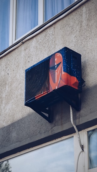 An air conditioning unit is mounted to the exterior of a building, creatively painted with artwork resembling a futuristic helmet. The building's textured concrete wall and nearby window are visible.