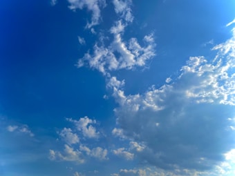 A clear sky filled with white, fluffy clouds scattered across a bright blue background. The clouds vary in size and shape, with some forming wispy patterns while others are more dense. Sunlight creates subtle variations in color depth and highlights the edges of the clouds.