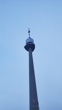 A tall communication tower extends into the sky, featuring multiple sections with a large circular platform near the top. Antennas and other technical equipment are visible at the pinnacle of the structure.