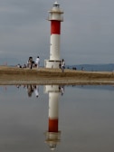 A lighthouse with a red and white pattern stands on a sandy beach, reflected in a shallow pool of water. People are walking and sitting around, creating a scene of leisure and relaxation. The sky is overcast, adding a sense of calm to the environment.