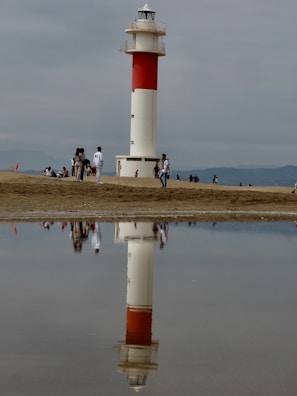 A lighthouse with a red and white pattern stands on a sandy beach, reflected in a shallow pool of water. People are walking and sitting around, creating a scene of leisure and relaxation. The sky is overcast, adding a sense of calm to the environment.