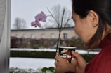 A person is holding a cup labeled 'Chocolat Noir' with a garden view in the background. An orchid with purple flowers is visible, and the environment appears to be wintery with snow covering the ground.