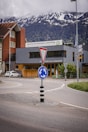 A roundabout with a directional road sign is situated in front of buildings, one of which is a hostel and coworking space. Snow-capped mountains are visible in the background under a cloudy sky.