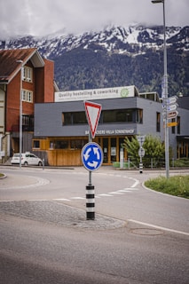 A roundabout with a directional road sign is situated in front of buildings, one of which is a hostel and coworking space. Snow-capped mountains are visible in the background under a cloudy sky.
