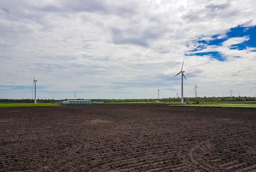 A wide expanse of agricultural land with neatly plowed soil dominates the foreground. Several wind turbines are scattered across the landscape, their tall white structures contrasting against the overcast sky with patches of clear blue. A line of trees and vegetation can be seen in the background, behind which stretches a long building complex with light-colored roofs.