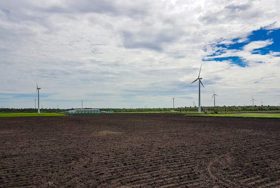 A wide expanse of agricultural land with neatly plowed soil dominates the foreground. Several wind turbines are scattered across the landscape, their tall white structures contrasting against the overcast sky with patches of clear blue. A line of trees and vegetation can be seen in the background, behind which stretches a long building complex with light-colored roofs.