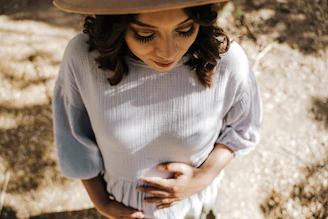 A person wearing a light-colored blouse and a hat is gently holding their stomach. The scene is sunlit, casting soft shadows and highlighting the serene expression on their face.