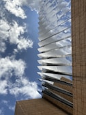 A modern architectural structure with reflective metallic elements extends upwards towards a blue sky dotted with clouds. The building's base is clad in beige stone, providing a contrast to the sleek metal lines.