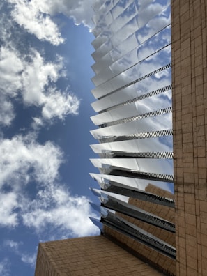A modern architectural structure with reflective metallic elements extends upwards towards a blue sky dotted with clouds. The building's base is clad in beige stone, providing a contrast to the sleek metal lines.
