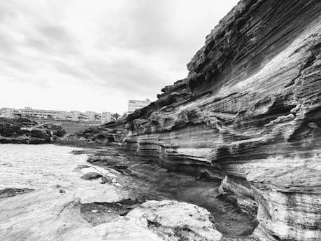 A dramatic coastline with rugged, layered rock formations extending alongside the ocean. The foreground shows intricate patterns in the rock, revealing geological history, while buildings are visible in the distance across a rocky shore. The sky is overcast, creating a somber atmosphere.