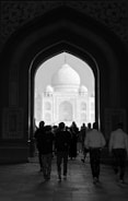 A group of people walks through an ornate archway towards a large, domed structure in the distance, which is the Taj Mahal. The intricate details of the architectural design frame the scene, and the pathway is dimly lit, creating a contrasting silhouette effect.