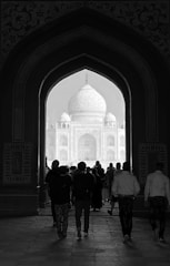 A group of people walks through an ornate archway towards a large, domed structure in the distance, which is the Taj Mahal. The intricate details of the architectural design frame the scene, and the pathway is dimly lit, creating a contrasting silhouette effect.