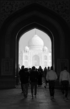 A group of people walks through an ornate archway towards a large, domed structure in the distance, which is the Taj Mahal. The intricate details of the architectural design frame the scene, and the pathway is dimly lit, creating a contrasting silhouette effect.