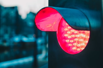 A close-up of a red traffic signal, with a focus on the illuminated lights beneath a metallic hood. The background appears blurred, suggesting a cityscape in soft focus with tones of blue and grey.