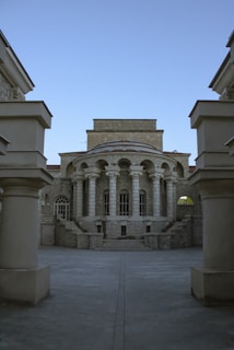 A classical stone building with a series of tall columns supporting an arched facade. The architecture is symmetrical with large windows and stone steps leading to the entrance. The structure is framed by two large pillars in the foreground, and the sky is clear and blue.