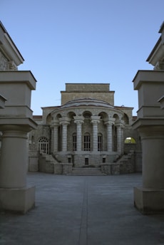 A classical stone building with a series of tall columns supporting an arched facade. The architecture is symmetrical with large windows and stone steps leading to the entrance. The structure is framed by two large pillars in the foreground, and the sky is clear and blue.