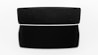 A black speaker with a sleek, modern design is centered against a white background. The speaker is rectangular with fine mesh detailing on the front. A shadow extends below it, accentuating its dimensions.