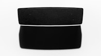 A black speaker with a sleek, modern design is centered against a white background. The speaker is rectangular with fine mesh detailing on the front. A shadow extends below it, accentuating its dimensions.
