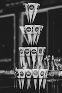 A black and white photograph displaying multiple tiers of conical paper wrappers, each likely used for holding snacks or small items. They are neatly arranged in a pyramid-like structure and appear to be labeled with different messages or flavors, such as 'HAVSSALT' and 'CHAMPAGNE JORDBUBB'. In the background, there are blurred glass bottles visible.