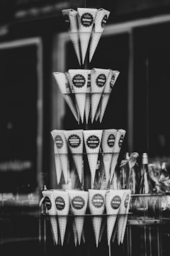 A black and white photograph displaying multiple tiers of conical paper wrappers, each likely used for holding snacks or small items. They are neatly arranged in a pyramid-like structure and appear to be labeled with different messages or flavors, such as 'HAVSSALT' and 'CHAMPAGNE JORDBUBB'. In the background, there are blurred glass bottles visible.