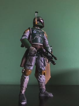 A detailed action figure of a character in armor stands against a green background. The figure is holding a blaster rifle, wearing a helmet with a visor and antenna, and has a cape draped over one shoulder. The armor features a mix of green, grey, and brown colors, with intricate detailing and battle-worn effects.