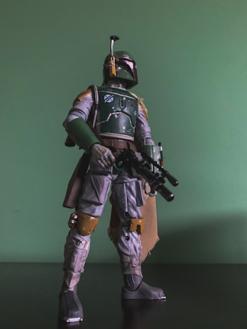 A detailed action figure of a character in armor stands against a green background. The figure is holding a blaster rifle, wearing a helmet with a visor and antenna, and has a cape draped over one shoulder. The armor features a mix of green, grey, and brown colors, with intricate detailing and battle-worn effects.