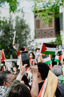 A group of people gathered outdoors, holding flags with red, black, white, and green colors. Many individuals are taking photographs or videos with their phones. The background shows greenery and a building with decorative wooden panels.
