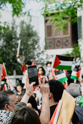 A group of people gathered outdoors, holding flags with red, black, white, and green colors. Many individuals are taking photographs or videos with their phones. The background shows greenery and a building with decorative wooden panels.